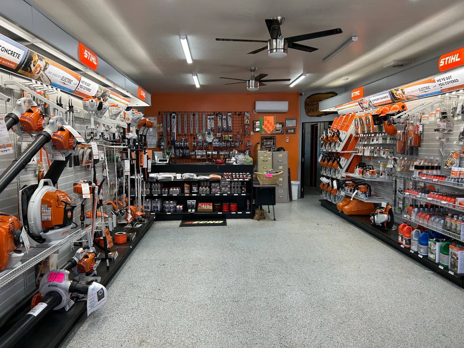 Interior of Woodchuck Saw Shop — orange walls, ceiling fans, STIHL chainsaw and accessories displays running down both sides of the aisle, ECHO floor mat